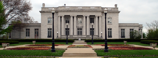 A pillared, two-story, gray marble building with several flower gardens in front