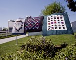 Three examples of Lancaster Amish Quilts shown outside hanging on a line in Lancaster, Pennsylvania. The patterns on these quilts are as follows: A heart made of a wreath of flowers on a white background with a blue block border (Left); Blue, Red, Purple, and green chevrons on a burgundy background (Middle); Depictions of Amish outfits including work shirts and suspenders, hats, pants, and hair coverings on a white background with a teal block border.