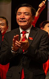 Bùi Văn Cường seen smiling and clapping, wearing a black suit, red tie and white shirt while having a National Assembly pin on his right chest.