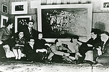 Several men and women are sitting and standing in front of a wall. This wall is decorated with many surreal art pieces.