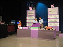 A television studio. Items are displayed on low tables, while in the back are two boards listing items and a sponsor logo each and topped with the letters W and K.