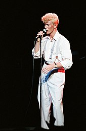 A long photograph of a man with bleached blonde hair and a white suit speaking into a microphone