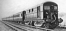 A black and white image of an electric locomotive hauling at least 6 coaches, shown with the electric locomotive on the right. There is an track in the foreground that is electrified with the fourth rail system, the locomotive is shown with two pickup shoes.