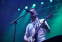 Frank Turner plays his electric guitar while singing into a microphone. The picture is taken from below, making it seem as if he is very high above the photographer.