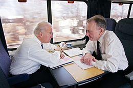 Two men are speaking to each other in a compartment on a train. Sitting in blue seats with paperwork spread out on tables in front of them, the man on the left has white hair, and the man on the right has gray hair. Both wear white dress shirts, black pants and ties.