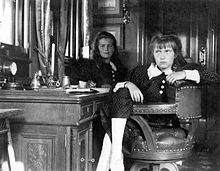 Maria, in background, holds a writing implement at the ready, while Anastasia sits jauntily on the desk's leather chair.