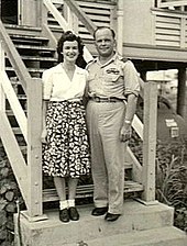 Informal full-length portrait of woman in skirt and blouse and man in light-coloured military uniform standing on steps in front of a building