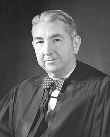 A black-and-white photographic portrait of an older man from the shoulders up, wearing judicial robes and a bow tie