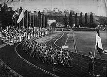 A black and white photo of a large track and field stadium with spectator stands and flags flying on the left hand side and trees in the background. A large procession of people in wheelchairs goes round the track with a sign being held at the front reading "Italia". The team is led by a walking man carrying the Italian flag.