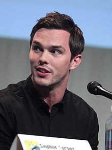A young, Caucasian man with short, dark hair and facial stubble wearing a black shirt speaks into a microphone against a grey background.