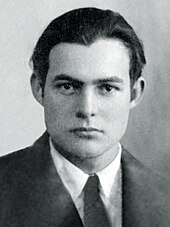 A black and white passport photograph of a young Ernest Hemingway wearing a dark suit and black tie. He is staring directly at the camera.