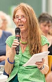 Eva Quistorp sitzt. Sie spricht in ein Mikrofon, das sie in der rechten Hand hält. In der linken Hand hält sie ein Schriftzug. Eva Quistorp hat schulterlanges rotes Haar, sie trägt eine rote Brille und eine grüne Bluse.