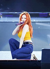 A red haired woman wearing a yellow top and blue jeans performing on stage while holding a microphone in her right hand.