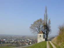 An old stone watch tower stands at a mountain top road; past the road, layers of rounded hilltops illustrate the kind of terrain the combatants faced. In the distance a silvery shimmer on the horizon marks the location of the Rhine river.