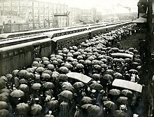 A large crowd of people, many of whom are holding umbrellas, are gathered at the train station to send off local recruits. Several factories can be seen behind the trains carrying the soldiers.