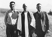 Three men stand in a field. The left-most is casually dressed and of aboriginal heritage, the middle one is a white man wearing ecclesiastical robes, and the right-most is a white man wearing a three piece suit.