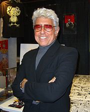 A portrait photograph of a man with white hair and sunglasses, smiling and crossing his arms.