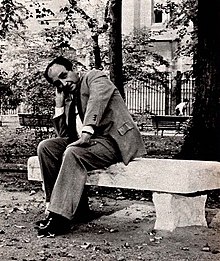 black-and-white photograph of Gatto dressed in a suit sitting on a bench and resting the side of his head on a hand while looking at the camera