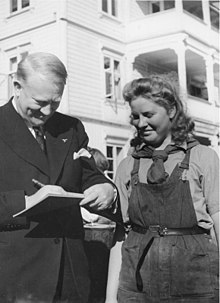 A middle-aged man in a dark coloured suit is writing on a pad of paper. Standing close to him and watching is a woman in her late twenties to early thirties. She is smiling, and wearing dark-coloured dungarees, with a shirt underneath and a scarf tied around her neck.