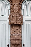 The highly decorated stoup in the porch of Landivisiau's Église Saint-Thuriau. This is fixed to the trumeau between the two entrance doors to the church. It is decorated with godrons (gadrooning) and beneath an elaborate canopy is the depiction of an angel holding a bottle brush or aspergillum ("Goupillon"), an allusion to the baptism. The renaissance dais features flat pilasters decorated with acanthus leaves, seashells and fire pots and the cartouches below depict four high relief carvings; the heads of two men, their feathered hats "à la Henri 111" alternating with the heads of two women whose long hair is held back by a hairband in the Renaissance style.