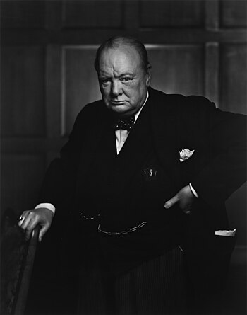 Black-and-white image of Winston Churchill standing with his left hand on his hip and his right hand resting on the back of a chair