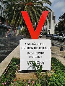 A letter V is placed on top of a pedestal. The pedestal has written the phrases "50 years after the state crime", "10 June 1971–2021" and "The struggle continues!" in Spanish.