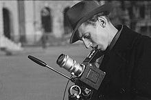 Seitliche Schwarzweißfotografie eines Mannes mit Hut, der von oben durch den Sucher blickt. Die Kamera steht auf einem Stativ und ist mit seinem langen Objektiv nach oben gerichtet.