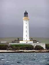A tall white lighthouse with a brown stripe around the parapet and dark coloured lantern sits on a rocky shore. A white wall obscures the lower floor of grey stone buildings gathered around its base.