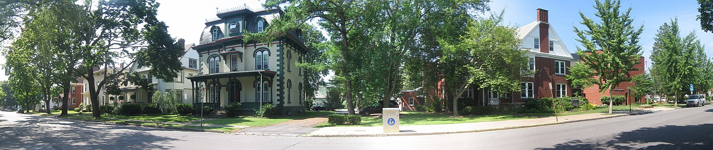 A tree-lined street with large houses in bright sunshine; at far right is a red brick building with a sign reading "Ross Library"