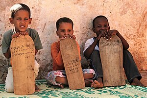 three young brown-skinned boys sit or kneel on a green patterned carpet against a plastered wall. They each hold a flat piece of wood about half their size. The pieces of wood are straight on 3 sides but curved at the top, with Arabic writing.