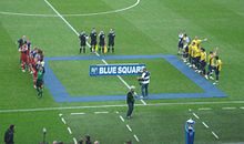 Two football teams line up opposite each other on the halfway line, with the referee, assistants and the fourth official standing to their side