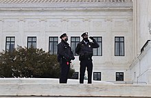 Two D.C. police officers wearing black face masks stand on top of a ledge in front of the U.S. Supreme Court building.