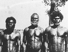 drei Aborigines verschiedenen Alters mit muskulösen Oberkörpern, welche mit ernstem Blick in die Kamera schauen
