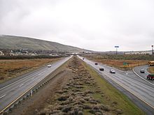 A four-lane freeway with a wide median approaching a residential neighborhood with an overpass in the distance.