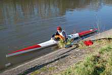 Long, thin kayak with blunt bow and stern, on flat water, person getting in
