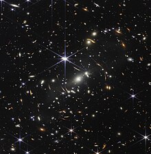 The background of space is black. Thousands of galaxies appear all across the view. Their shapes and colors vary. Some are various shades of orange, others are white. Most stars appear blue, and are sometimes as large as more distant galaxies that appear next to them. A very bright star is just above and left of center. It has eight long, bright-blue diffraction spikes. Between 4 o'clock and 6 o'clock in its spikes are several very bright galaxies. A group of three are in the middle, and two are closer to 4 o'clock. These galaxies are part of the galaxy cluster SMACS 0723, and they are warping the appearances of galaxies seen around them. Long orange arcs appear at left and right toward the center.