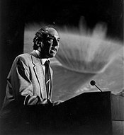 An elderly man with descending hair talking behind a speaker's desk with a microphone. There is a picture of an explosion on the surface of a planet in the background.