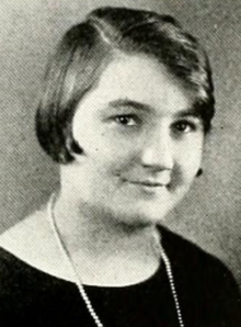 A young white woman with dark bobbed hair, parted at the side, wearing a black top with a round neckline, and a strand of pearls