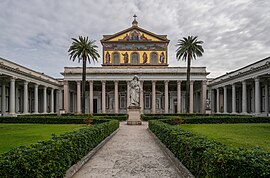 A courtyard with palm trees and a greater-than-lifesized statue of Saint Paul holding a sword in front of the colossal portico of the basilica and a large mural covering the upper facade