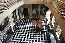 A photograph looking down on the interior of the Great Hall, showing the chequerboard floor tiles, several of the large wall portraits and pieces of furniture placed at the edges of the room.