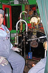 A smiling fireman sitting inside the locomotive's cab next to an assortment of valves, levers, and gauges