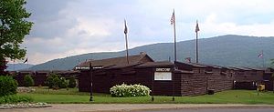 The reconstructed fort is a wooden log construction, painted brown, roughly a single story tall. There are four flagpoles, from which fly a variety of flags, including the American flag and a British Union Jack. Mountains are visible in the background.