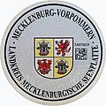 Aktuelle Zulassungs­plakette des Land­kreises Mecklenbur­gische Seenplatte mit dem mecklenburg-vorpommerschen Landeswappen, rechts mit DataMatrix-Code und Landkreiswappen oben links