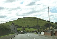 two-lane road bending downhill and to the right with a conical green hill beyond under a bright, cloudy sky