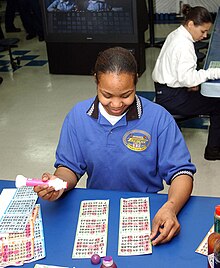 A Navy woman, seated at a table, has an assortment of bingo cards in front of her.