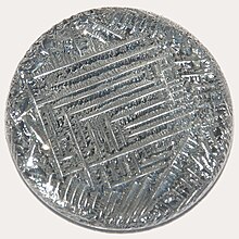 A shiny silver-white medallion with a striated surface, irregular around the outside, with a square spiral-like pattern in the middle.
