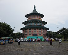 Bild einer chinesischen Pagode, vor der ein großer gepflasterter Platz ist, auf dem Autos und Menschen stehen.