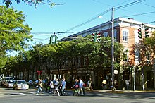 A street corner in a developed urban area with two-story ornate brick buildings and busy tree-line sidewalks under a blue sky. Traffic lights governing the intersection are red.