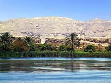 In the foreground the Nile, in the middle ground, luxurious plants and palms trees, in the background the barren hills of the desert