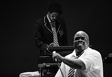 Two African-American men perform against a black backdrop; the man in the background plays a keyboard whilst wearing a striped tie and sunglasses, whilst the man in the foreground holds a microphone whilst wearing a white shirt and striped tie.